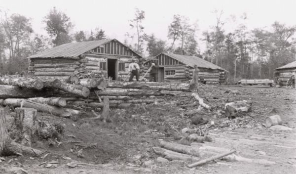 Madeline Island logging camp near Big Bay. Men preparing logs for a building, with three log buildings in background.