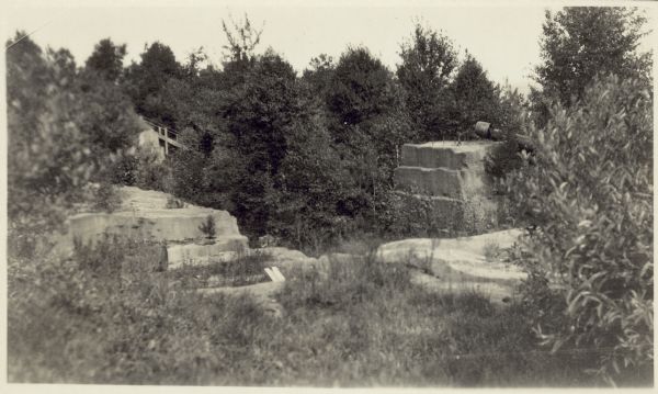 Quarry at Hermit Island (Wilson Island). Stones, plants and a foundation are in the foreground. In the background among trees is a wooden structure.