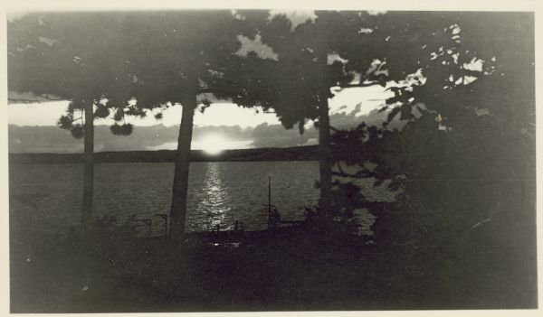 View through trees of sunset, probably over Lake Superior. A forested shoreline is in the far background across the water.