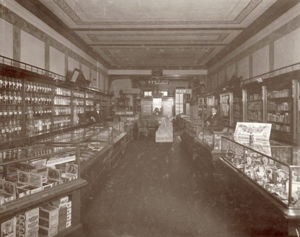 Interior view of Trukenbrod's Pharmacy. The signs in the rear of the store proclaim that they are proprietors of both "Prescriptions" and "Paints" alike, which was very common for the time period. A Johann Hoff's Malt Extract display testifies to their probable sizable stocks of proprietary medicines. There are men is standing behind glass cases on the left and right. On the immediate left is a glass case and shelf displaying cigars.
