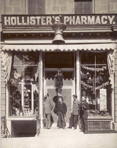 Exterior view of A.H. Hollister's Pharmacy, which was located at 3 North Pinckney Street. The man with the long beard (far left), is Albert H. Hollister (1843-1910). The young man to his right is Frank Swain. The other two men are unidentified.