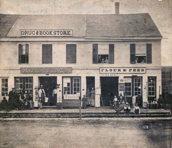 Street scene with customers in front of the M.P. Roberts Drugstore and C. Baack Flour and Feed Store.