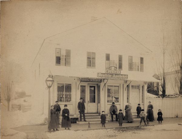 Charles Pfeifer stands on the entrance steps to the drugstore with his wife Emma Pfeifer, who holds a baby. Other children and adults pose with them.