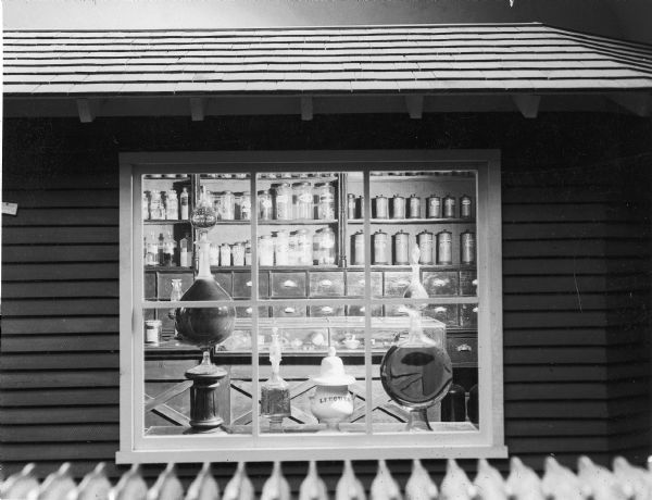Interior view of the Wisconsin Historical Society Pioneer Drugstore Exhibit. Pictured are several decorative showglobes, a leech jar, and shelves of various pharmaceutical jars.