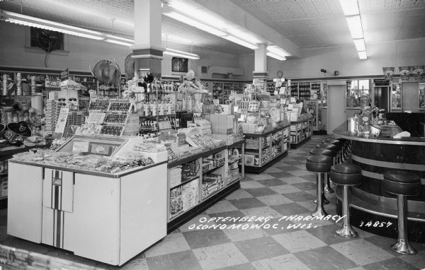 Interior view of the Optenberg Pharmacy. On the left is a large candy counter, including a Russell Stover Candies display. A curved soda fountain counter is on the right.