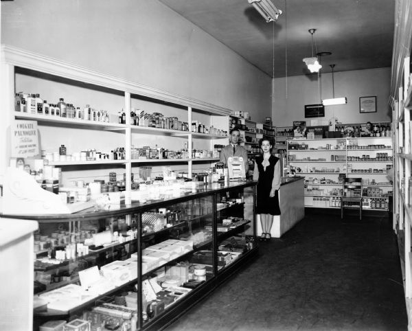 Thomas Lackland, proprietor of Lackland Pharmacy, poses with a clerk behind a well-stocked counter, full of a variety of personal hygiene items.