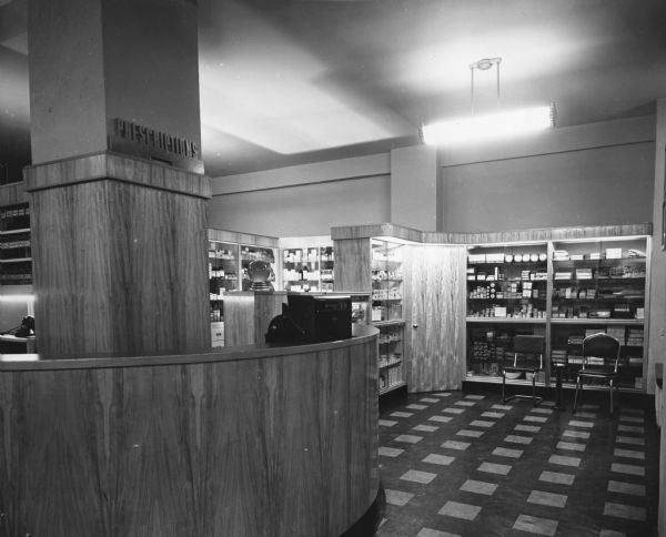 Image of a curved wooden counter, complete with cash register and traditional pharmaceutical showglobe, where customers placed and picked up their prescription orders. A small waiting area is on the right.