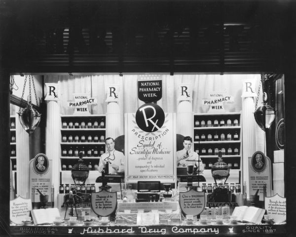 A window display at Hubbard Drug Company for National Pharmacy Week. The display extols the virtues of your pharmacist being able to craft medicines specifically for each consumer. Bottles of traditional medicines, a large mortar and pestle, as well as 4 showglobes are incorporated in the display.