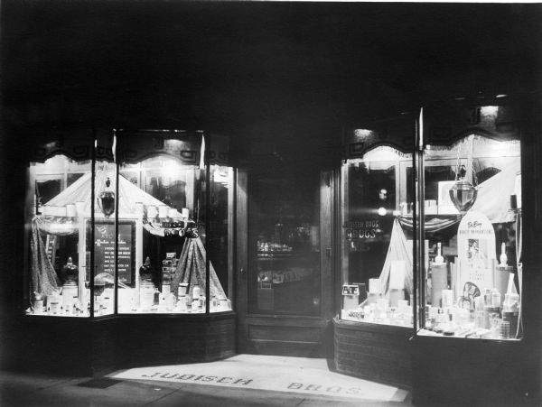 Decorative window displays in the storefront of Judisch Brothers' Drug Company. A variety of pharmaceutical glassware; mortars and pestles; and traditional show globes are utilized in the exhibit.