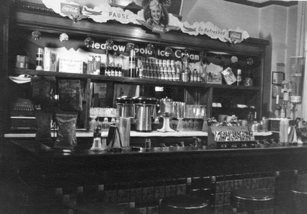 View of the soda fountain in Meyer's Pharmacy in Topeka. Advertisement premiums for Meadow Gold Ice Cream and Coca-Cola are featured prominently.