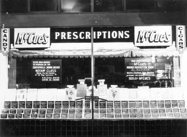 Exterior view of McCue's Drugstore. In the window are drawings of drug specimens produced by a local botany class.