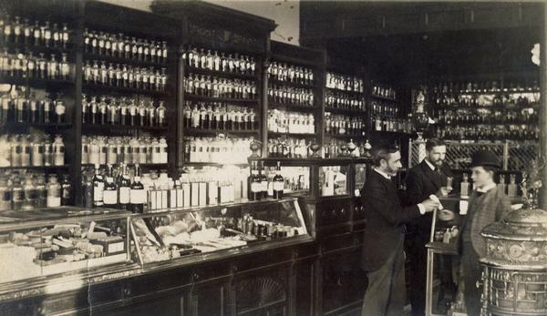 View of scene at J.N. Hegeman Drugstore, 1218 Broadway Street, New York. John Ferris hands a prescription written on a piece of paper to the errand boy. Frank Parker stands  at his left. The pharmacy had an impressive selection of medicinal liquors, but lacked the patent medicines that most other pharmacies stocked at the time.