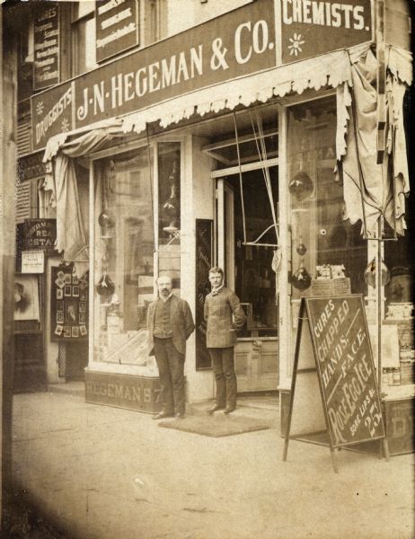 Joseph Glenny (left) and a clerk stand outside of the 756 Broadway branch of the J.N. Hegeman & Co. Drugstore. The front windows contain an impressive assortment of traditional pharmaceutical showglobes.