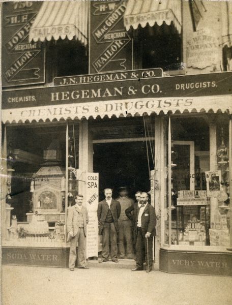 Exterior view of J.N. Hegeman & Company Drugstore. Pictured from right are proprietor J.N. Hegeman (with cane), Joseph B. Glenny, and three associates. The sign on the left details the assortment of spa waters the establishment carried, including the well-known cure-all, Vichy Water.