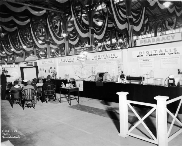 View of a U.S. Pharmacopeia Exhibit about a class of medications called 'digitalis' at the annual Philadelphia on Parade. Digitalis medicines are used to improve the strength and efficiency of the heart, or to control the rate and rhythm of the heartbeat. A man is standing on the left side of the exhibit giving a presentation to a small group of onlookers.