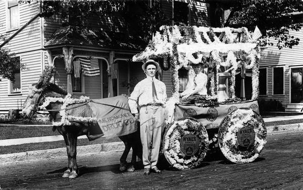 Rial Herreman stands beside a horse-drawn wagon/float, decorated with fresh flowers, advertising the Menomonee Drug Company. This may have been part of a local parade.