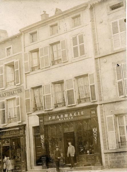 View of the exterior of the Pharmacie Werner in Boulay/Bolchen, France. This region was part of Germany in 1889. Two men are standing in front of the building.