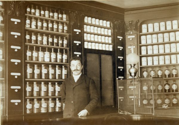 A pharmacist is standing in front of a case of labeled medical ingredients in the Pharmacie Werner in Boulay, France. This region was part of Germany when this photograph was taken.