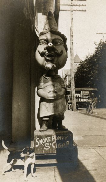 A small dog stands beside a large statue (appears to be carved out of wood) advertising Pompy Cigars, outside of a drugstore.