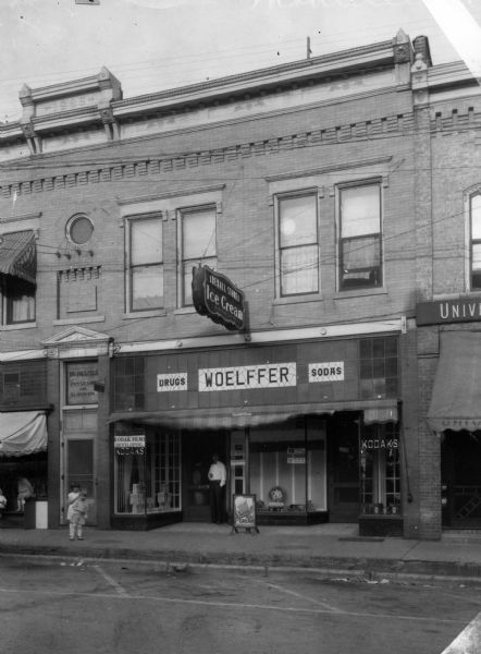 View from street towards Woelffer's Drug Store. Children are on the sidewalk, and a man is standing in the doorway. The store had a soda fountain with ice cream, and a film processing department.