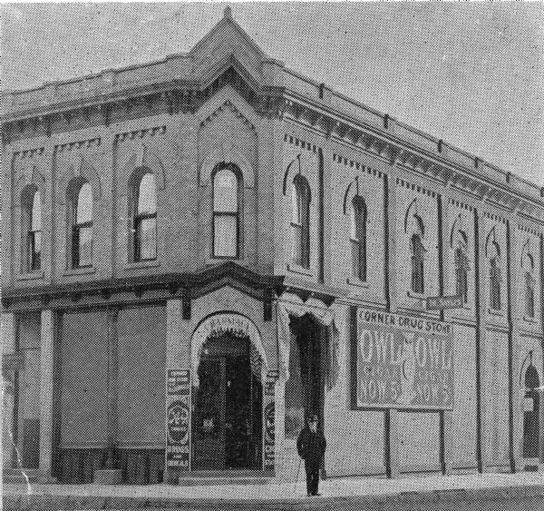 A man stands outside the doorway of The Corner Drug Store, corner of 3rd and Oak Streets.  On the side of the building is a large advertisement for Owl & Owl cigars.