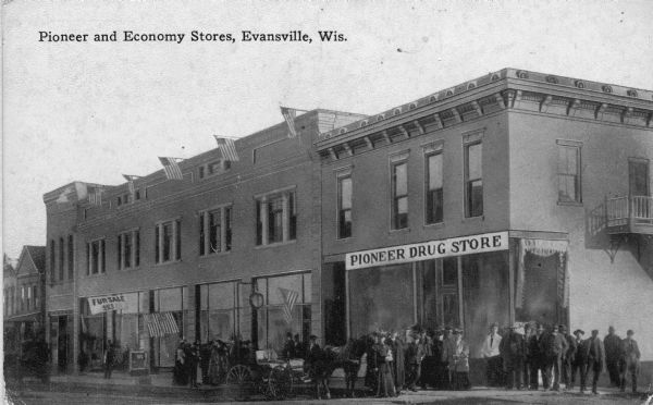 View from street towards people standing outside the entrance to the Pioneer Drug and Economy Store. Caption reads: "Pioneer Economy Stores, Evansville, Wis."