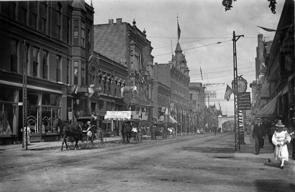 View of pedestrians and horse-drawn carriages downtown, at the corner of Main and Fifth Street. J.A. Erhart & Sons' Drugstore are on the right side of the road. L.G. Loomis' Music & Art store is directly across the street. The business sign is painted on a large elephant statue.