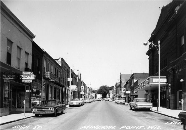 View of businesses on High Street, including the Rexall Drugstore on the left corner. One can also see a glimpse of the City Hall, directly across the street.