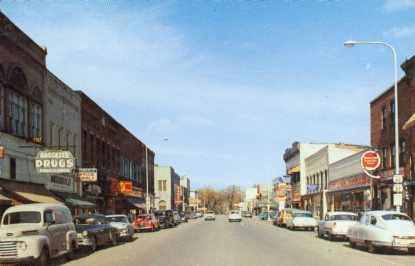 View of cars parked alongside businesses on Third Street. Bassett's Drugstore is on the left side of the street.