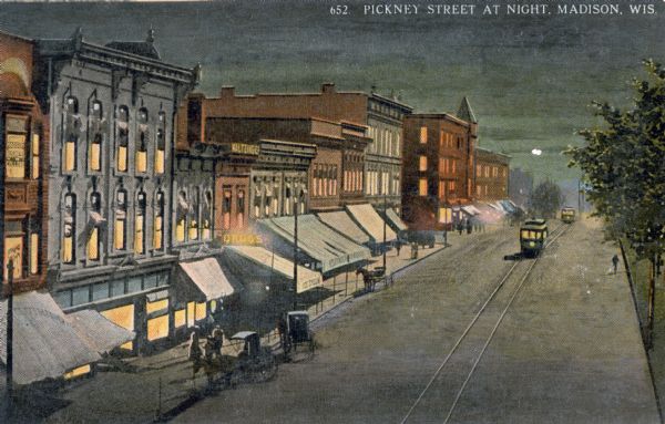 Elevated view at night of streetcars moving along the Capitol Square on Pinckney Street. Walzinger's Drugstore is one of the prominent storefronts on the left. Caption reads: "Pinckney Street at Night, Madison, Wis."
