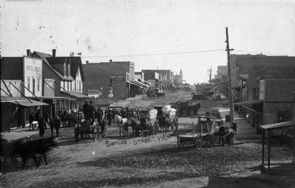 View of horse-drawn traffic in downtown Shattuck. Businesses on the left side of the street include the "White House," a saloon, and the Davis Drug company.