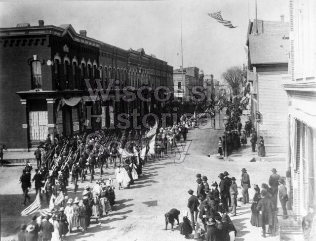 View of a large Columbus Day parade from Jackson and Third Streets. In the extreme right foreground is Conrad Althen's Store, and the Anderson and Larson Saloon is just behind it on the corner. Across the street is a large drugstore.