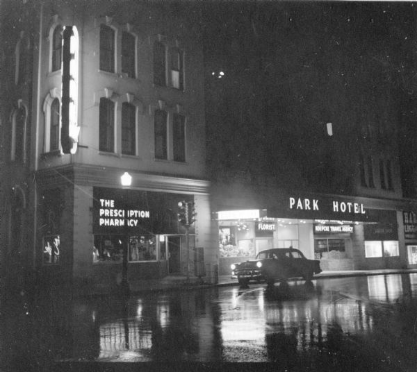 A rainy, nighttime view of the corner of Main and Carroll streets on the Capitol Square. The neon signs of the Prescription Pharmacy and the Park Hotel light up the wet pavement.