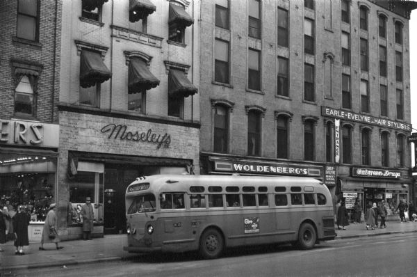 Bus traveling down Mifflin Street on Capitol Square. Moseleys, Woldenbergs and Walgreen's Drugs are on the right.