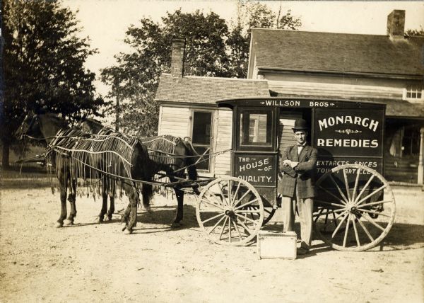 A traveling salesman with Willson's Monarch Labs is standing beside the horse-drawn wagon that took him on sales routes in rural Wisconsin. Willson manufactured patent medicines, extracts, spices, items for farm animals and toilet articles. The horses are wearing fly-nets.