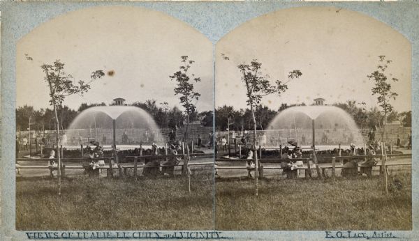 Stereograph of the Artesian Well near the intersection of Wisconsin and Minnesota (later renamed Wacouta) Streets. The following is printed on the backside of the photograph:
"The Greatest Artesian Well in America.
The Largest Flow in the World, of the Same Size Bore, with One Exception.
This well was completed in February 1876, bore 5 5/8 inches; pipe projects 10 feet above the ground the water falling into a basin 30 feet in diameter. A movable cap on top of the pipe obstructing the upward flow, and the water is forced outward and upward in all directions, by a pressure of 20,000 pounds.
Depth 960 feet or 970 from top of pipe. Temperature 56 F. Water flowed to top of pipe at a depth of 268 feet. Discharge per minute 603 9-10 gallons or nearly 30,000 bbls. Water will rise in a pipe 60 feet above ground or 100 feet above the Mississippi river. The whole distance that the water will rise is 1,020 feet above the bottom of the well. The pipe goes down 147 feet through sand and gravel to rock, first lime-rock 2 feet, shale 107 feet, sand rock 118 feet, sandy shale 90 feet, red and yellow ocher 6 feet, sandy shale 25 feet, sand-rock 4 feet (here brine begins to flow), sandy shale 71 feet, red, white and yellow sandstone in alternate layers, 355 feet, conglomerate of water-worn quartz-pebbles 35 feet, to bottom of well; which is 327 1/2 feet below the level of the ocean.
The water from this well is found to possess rare medicinal properties, and has cured many diseases of the bladder and kidney, Rheumatism, Dispepsia, St. Vitus dance chronic female complaints, and has infused new life and vigor into the aged and debilitated."