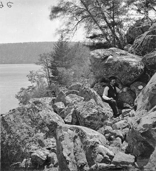 Devil's Lake Vicinity; "Effects of photographing among the rocks", foot of West Bluff. Man posing sitting and eating.