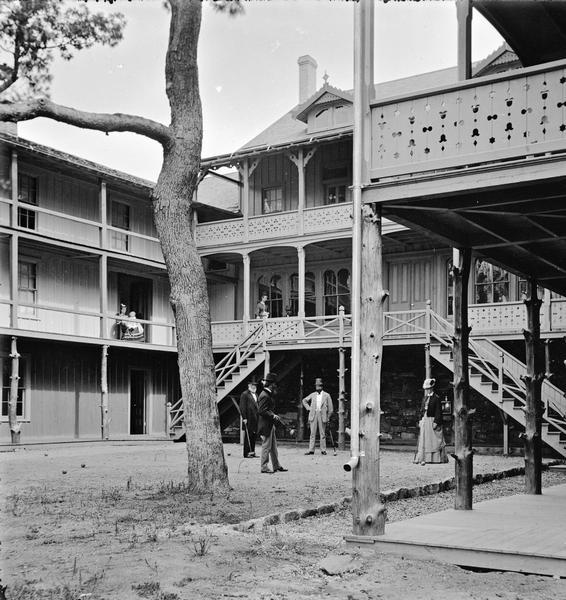 Devil's Lake vicinity. View of the Cliff House courtyard and croquet court. Three men and one woman are playing croquet. Two women are on the second floor balcony: one is walking at right, one is sitting (with child?) at left.