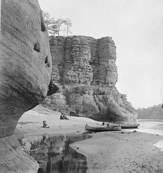 North face of High Rock. Two women are sitting on the sandy beach, one child is playing along the shoreline near a rowboat and canoe pulled up on the sandy shoreline, and another child is playing in the sand on the left. A man is standing partway up the face of High Rock on a ledge in the background.