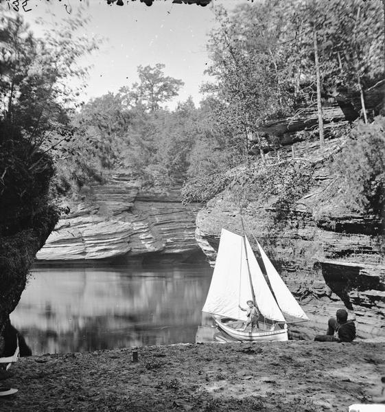 View out of Gates Ravine (Glen Eyrie). One man is reclining on the beach at right, and a second man is standing on a sailboat.