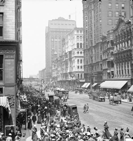 Elevated view of crowd along sidewalks on State Street, and the Masonic Temple from Madison Street. Streetcars and horse-drawn vehicles are in the street.