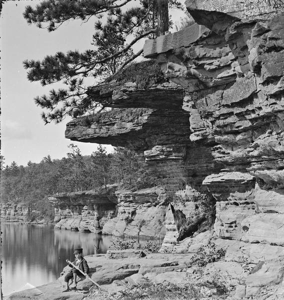 Up river from Angel Rock. There is a boy sitting on a rock, holding a pole, with rock ledges overhead.