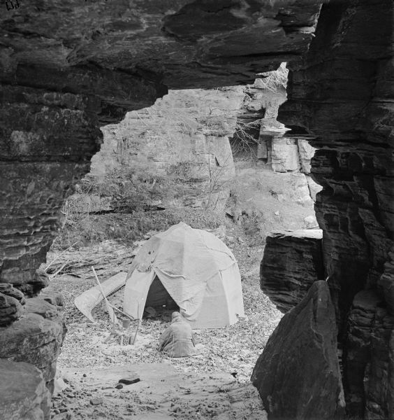 View through rock arch towards a man sitting on the ground facing away towards a tent and canoe, at the mouth of Chapel Gorge, foot of the Narrows.