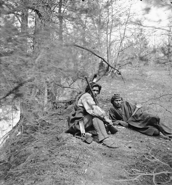 Coon-Nu-Gah (First Boy) and Big Bear, two Ho-Chunk men sitting on the ground. The man on the left is holding a gun.