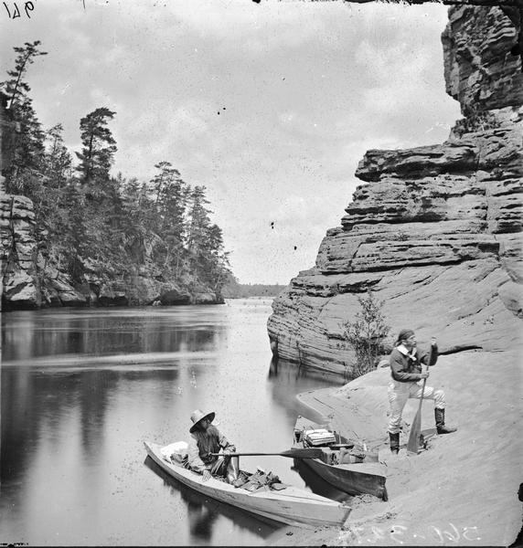 High Rock; the base with two rowboats. One man is sitting in a rowboat, and one man is standing on rocks and sand holding an oar.