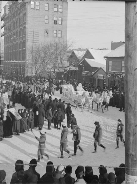 Elevated view of the St. Paul Ice Carnival; royal family equipages in street parade with crowd lining street. There is a real estate agency and a laundry on the right, and power lines are overhead.