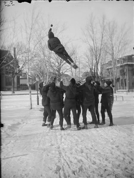 St. Paul Ice Carnival, bouncing — an instantaneous view. Circle of men in a residential area tossing a person in the air.