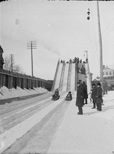 St. Paul Ice Carnival, Toboggan Slide in Palace Grounds. View looking up track towards people grouped to the side of toboggan tracks, on stairs, and at the top of slide. Two toboggans with people riding it are at the bottom of the slide. Power lines are overhead, and buildings are behind the slide.