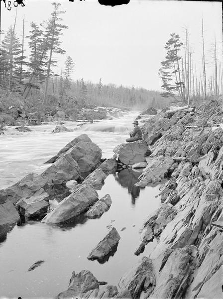 Dells of the St. Louis River, below the Caldron. A man is sitting on rocks at the edge of the river.