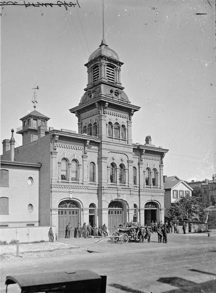 View across street towards the Milwaukee Central Fire Station, with fire fighters posing in front of the building and around a fire engine.